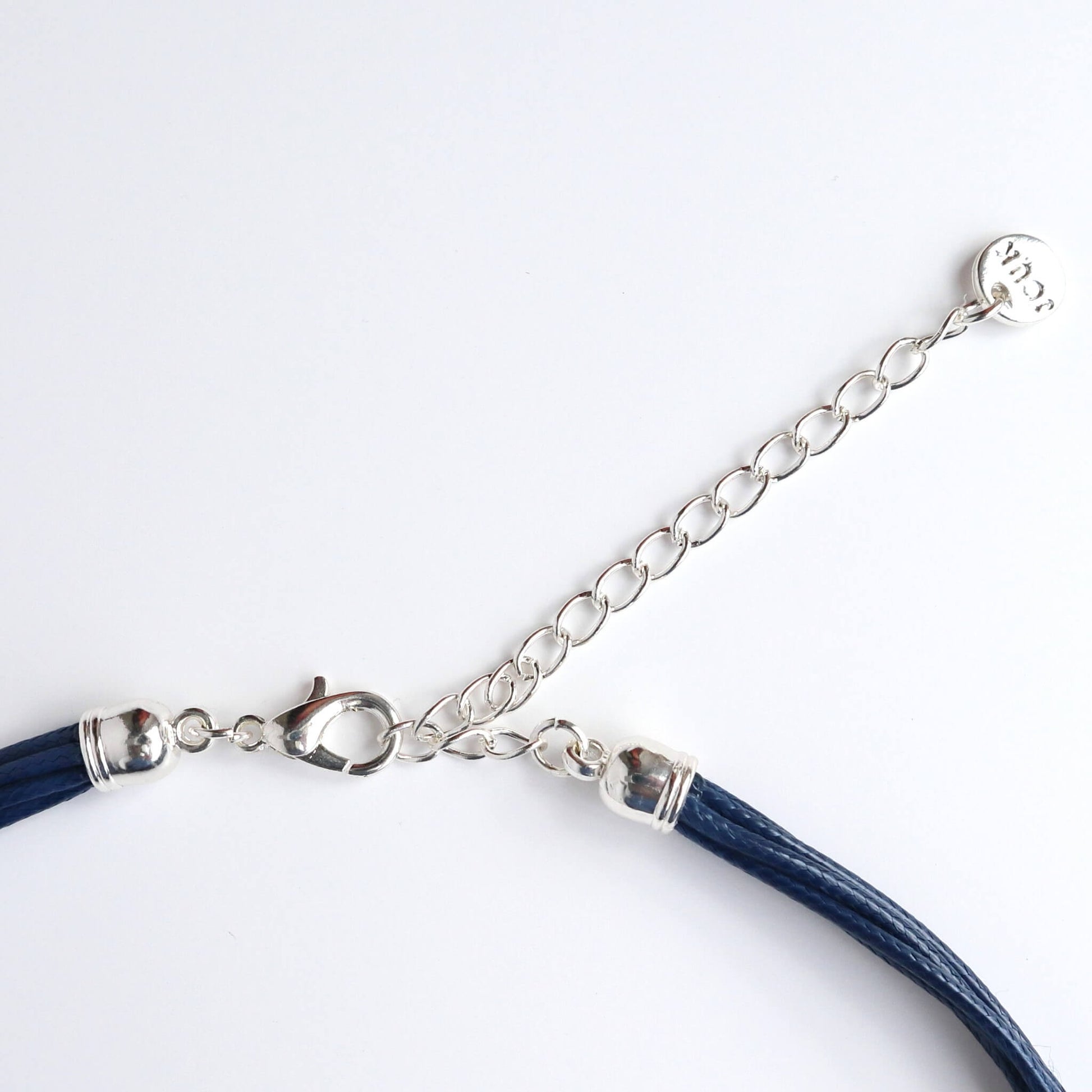 Close-up of a silver-toned statement necklace clasp and extender chain. Features a lobster claw closure and a small charm with lettering. Navy blue cords are visible.