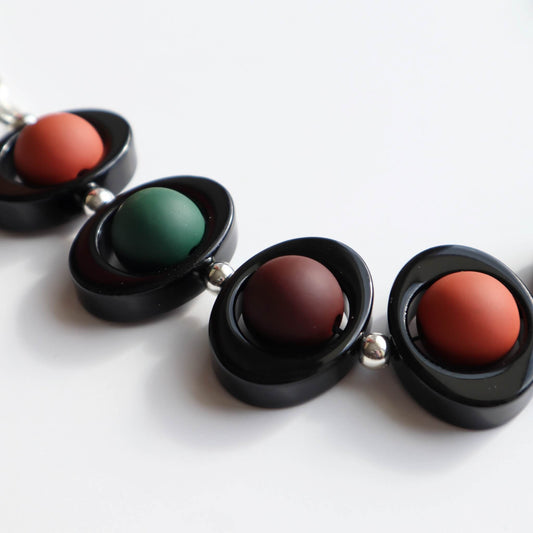 Close-up of a chunky necklace featuring black oval links with matte burnt orange, dark green, and deep red circular beads. Silver-toned chain visible.