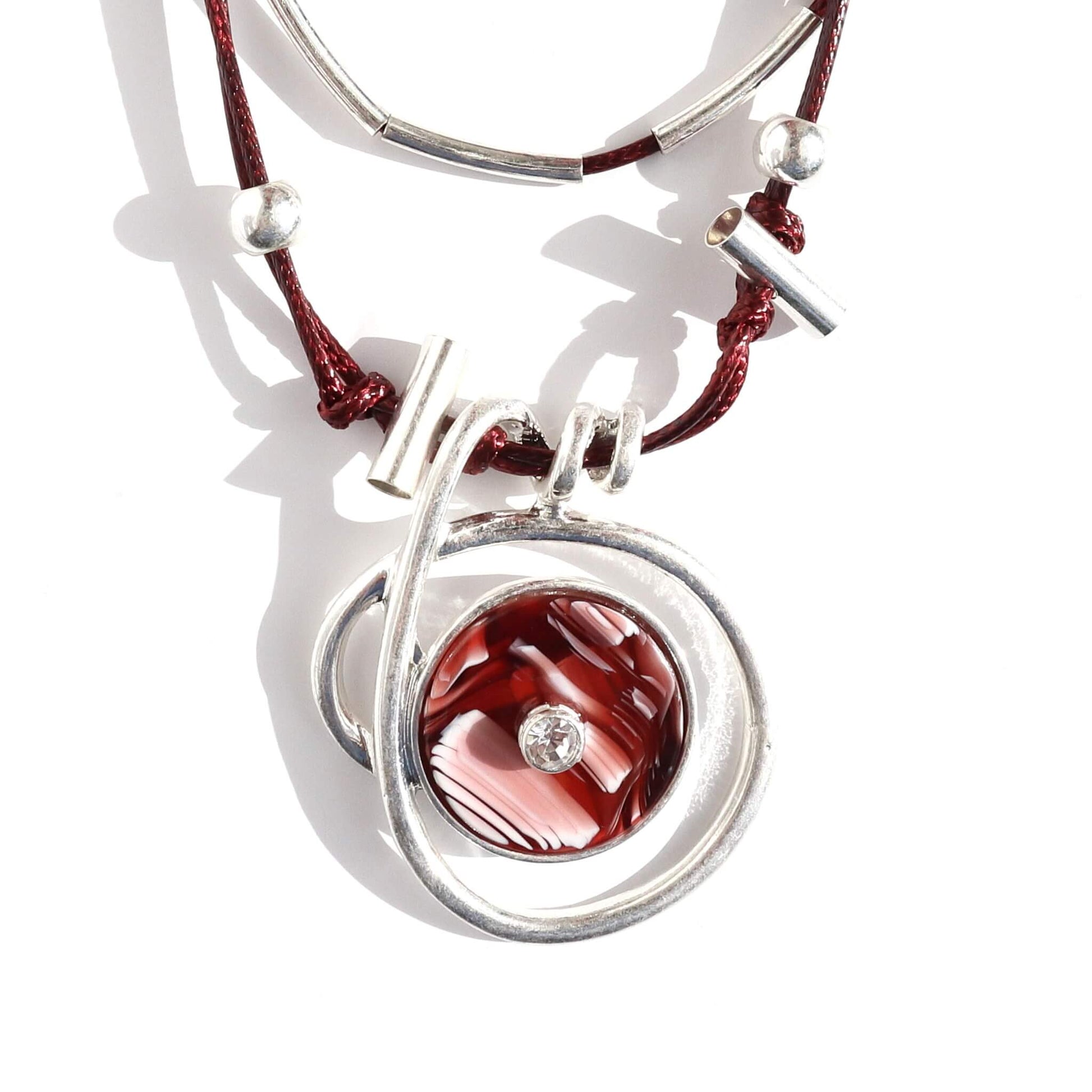 Detailed view of a layered statement necklace with deep red cords and silver-tone accents. Features a circular pendant with a red and white marbled centre and a small clear stone.