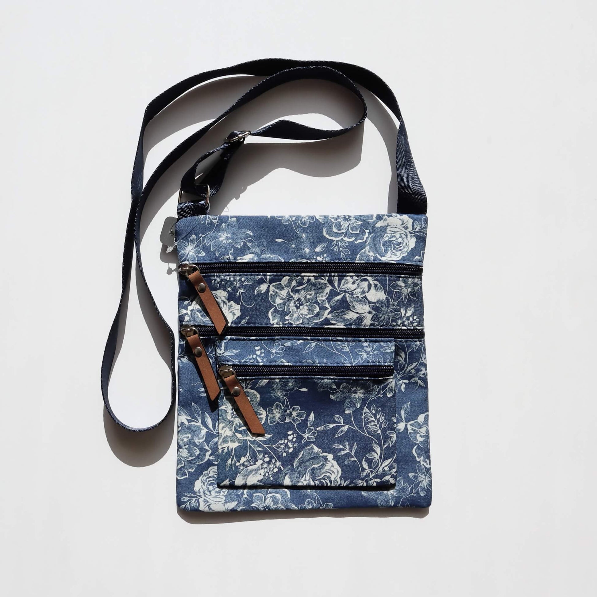 Dark denim crossbody bag with a white floral pattern. Features three zippered compartments with brown faux leather pulls and a dark blue adjustable strap.