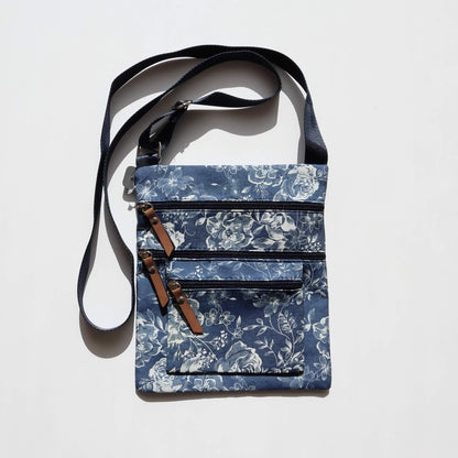 Dark denim crossbody bag with a white floral pattern. Features three zippered compartments with brown faux leather pulls and a dark blue adjustable strap.