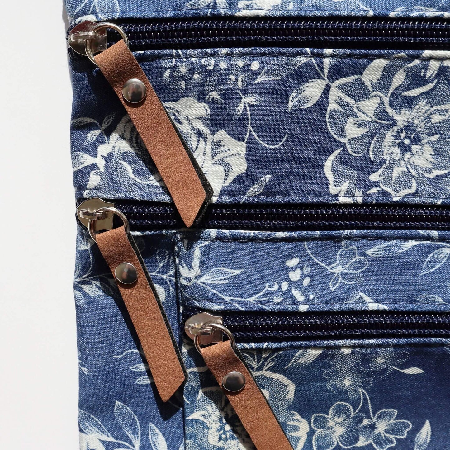 Close-up of a dark blue floral crossbody bag showing three zippered compartments with silver pulls and brown faux leather tabs.