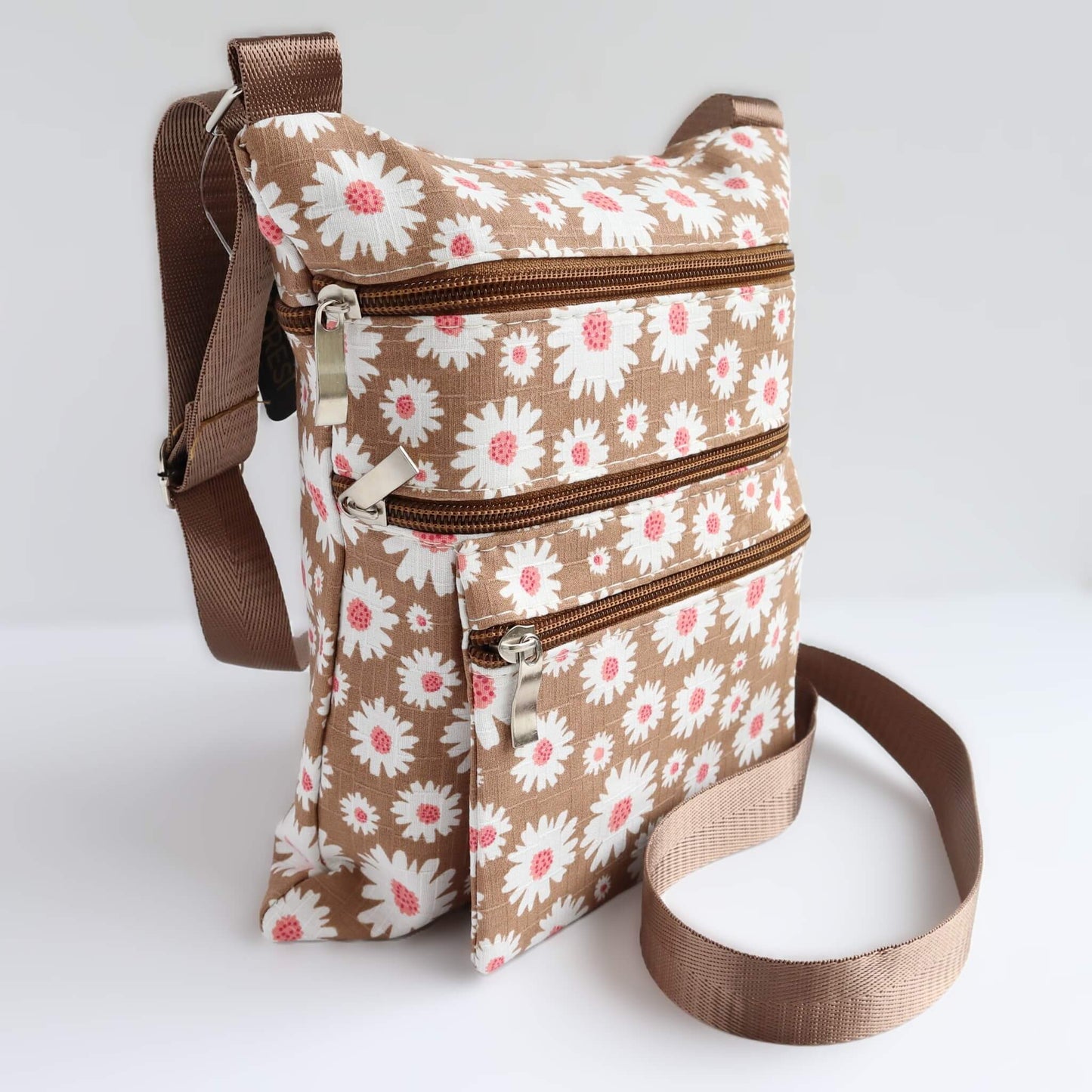 Compact brown bag with a daisy print in white and pink. Features three zippered sections and a brown, adjustable crossbody strap.