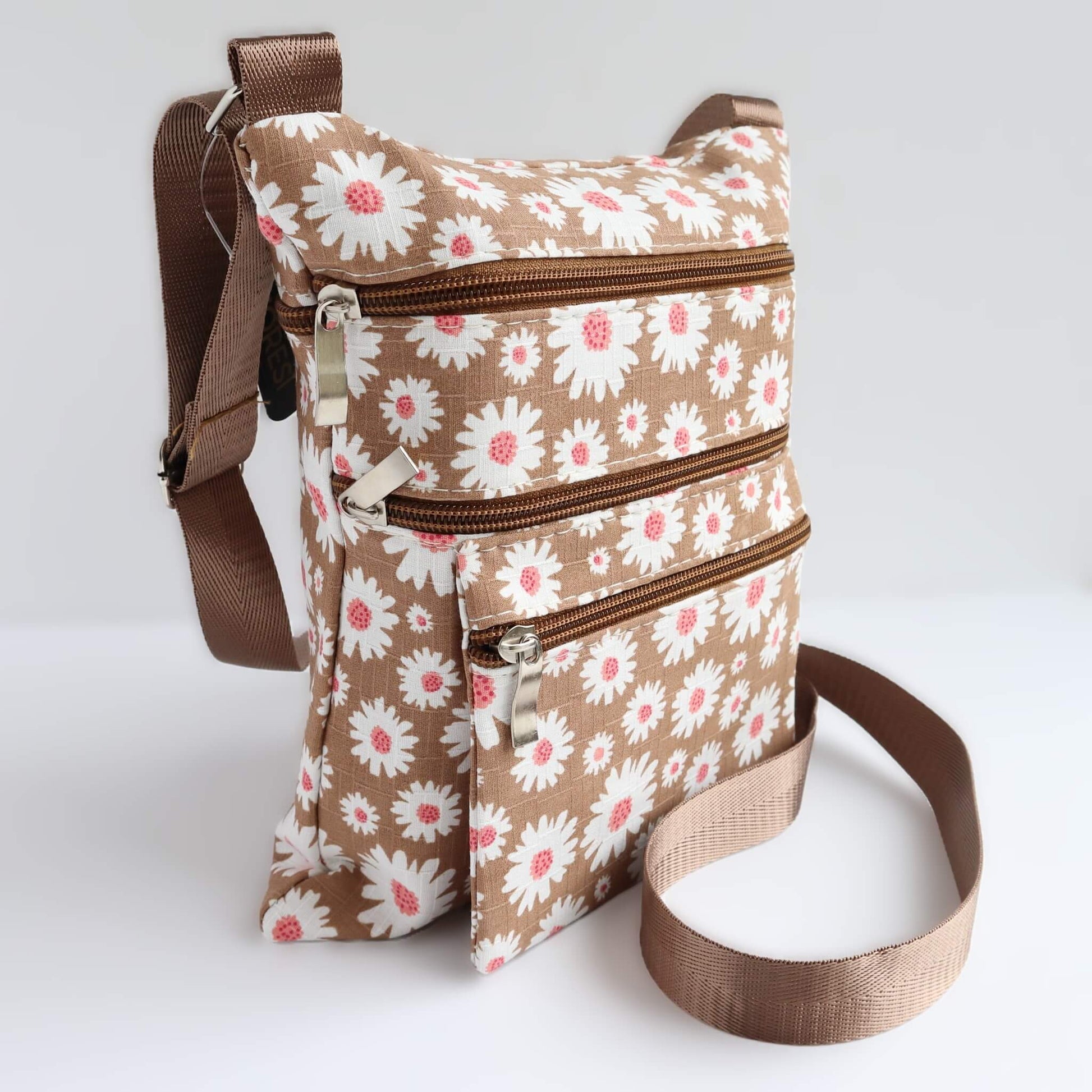 Compact brown bag with a daisy print in white and pink. Features three zippered sections and a brown, adjustable crossbody strap.