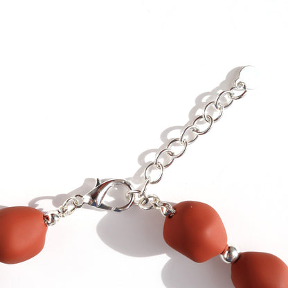 Close-up of a silver-toned necklace clasp and extender chain. Features a lobster claw closure. Matte rust-orange beads and a small silver spacer are visible.