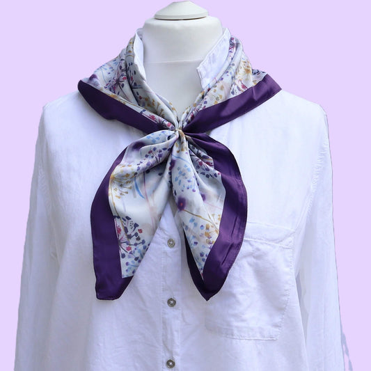  Aurora's Array: The Purple Dandelion Satin Square Scarf - A light satin scarf with a delicate dandelion and wildflower print in shades of purple, blue, and white, bordered by a deep purple satin edge, tied neatly around the neck of a mannequin wearing a 