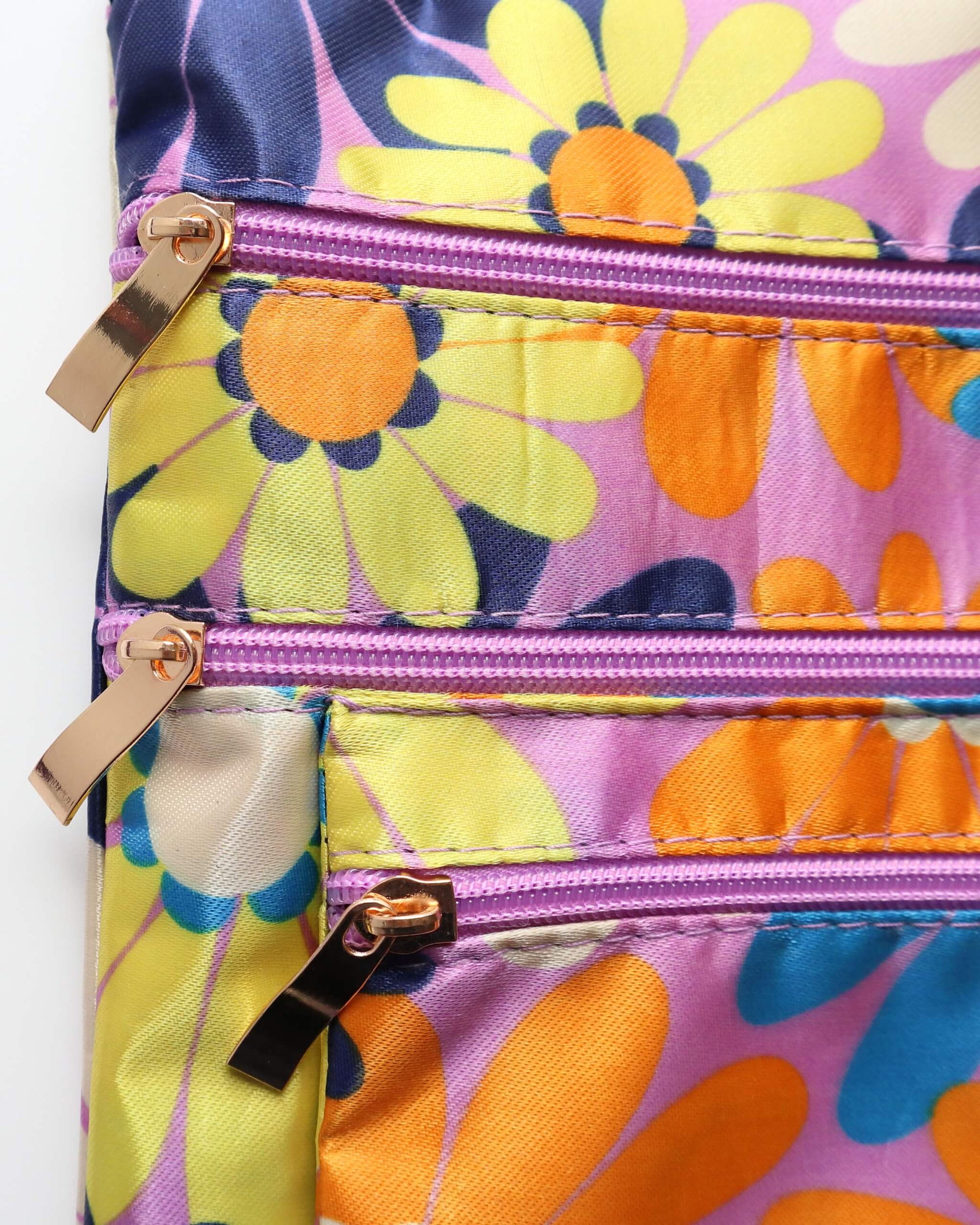 Close-up of a colorful crossbody bag with three zippered compartments featuring gold-toned pulls and purple accents. Vibrant daisy floral pattern.