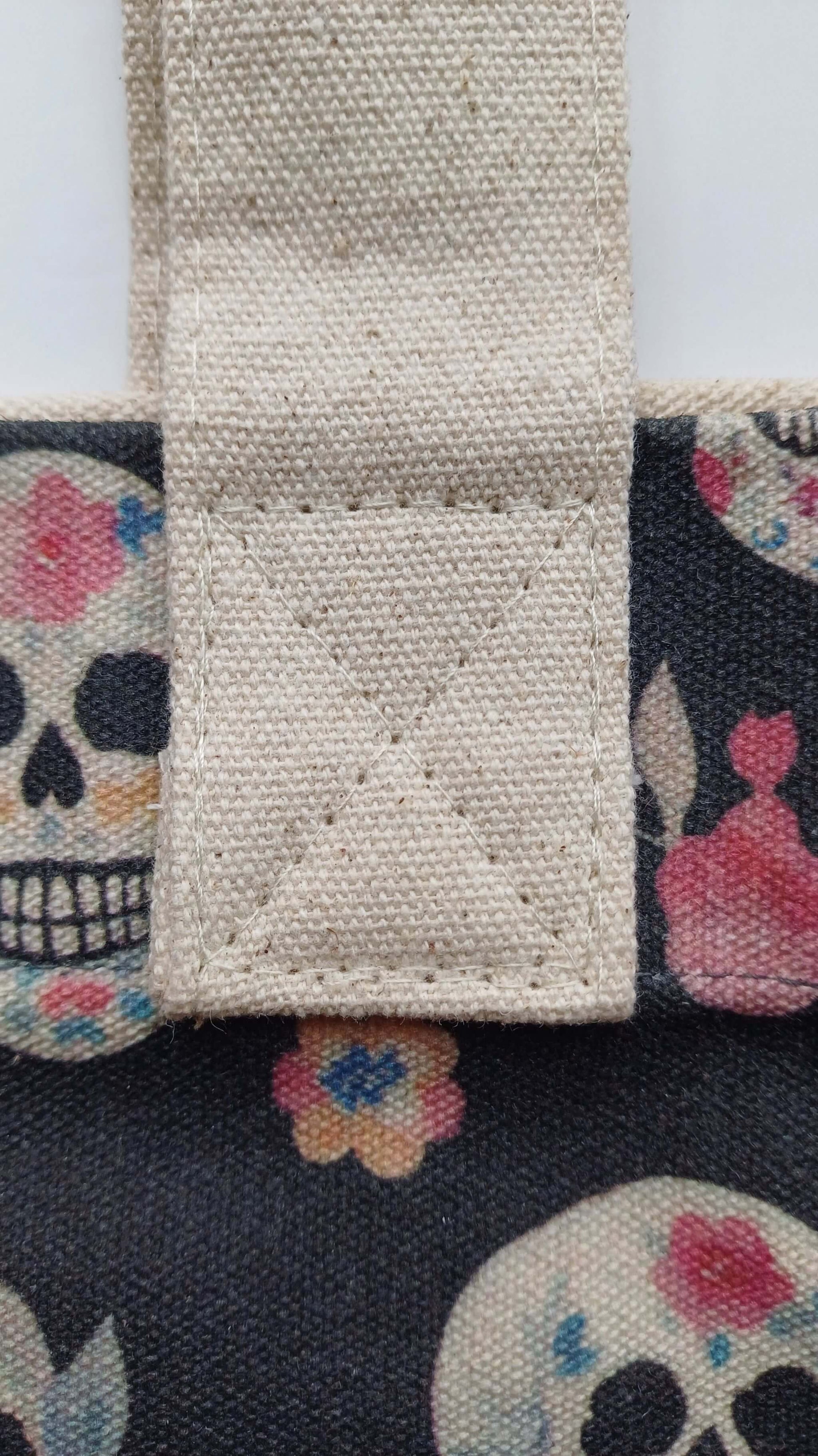 Close-up of a cream-colored canvas strap attached to a dark grey tote bag with a skull and floral pattern. Reinforced stitching detail.