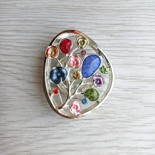 A decorative oval brooch featuring a silver-toned tree design filled with colorful enamel flowers and leaves, photographed on a light wood surface.